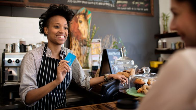 A woman working in a café holds a bank card as she speaks to a customer